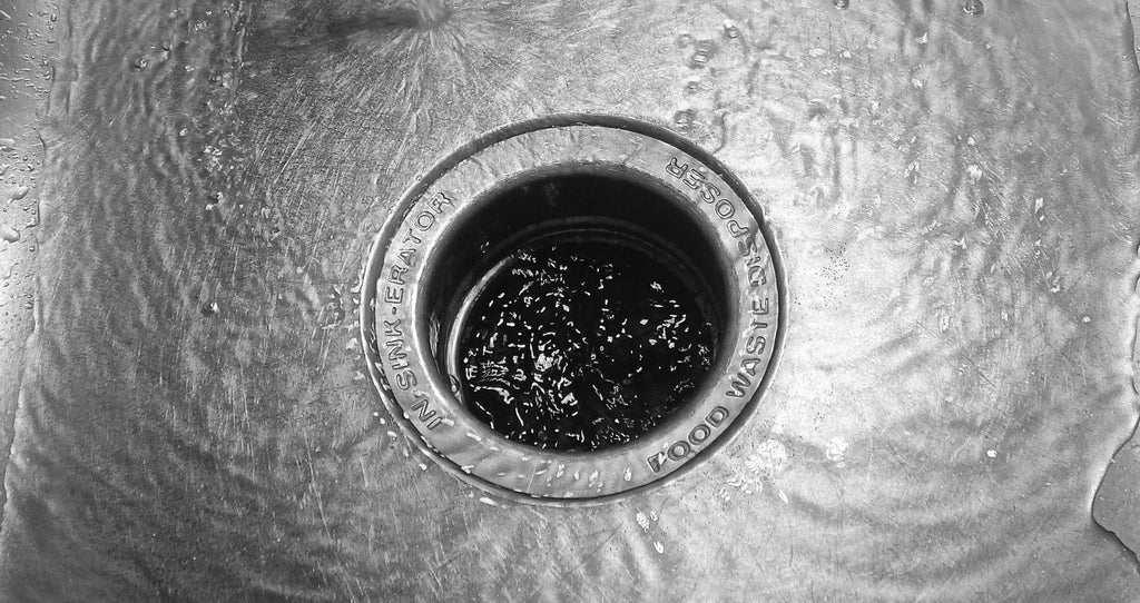 Water running into a stainless steel sink drain with food debris visible in the opening