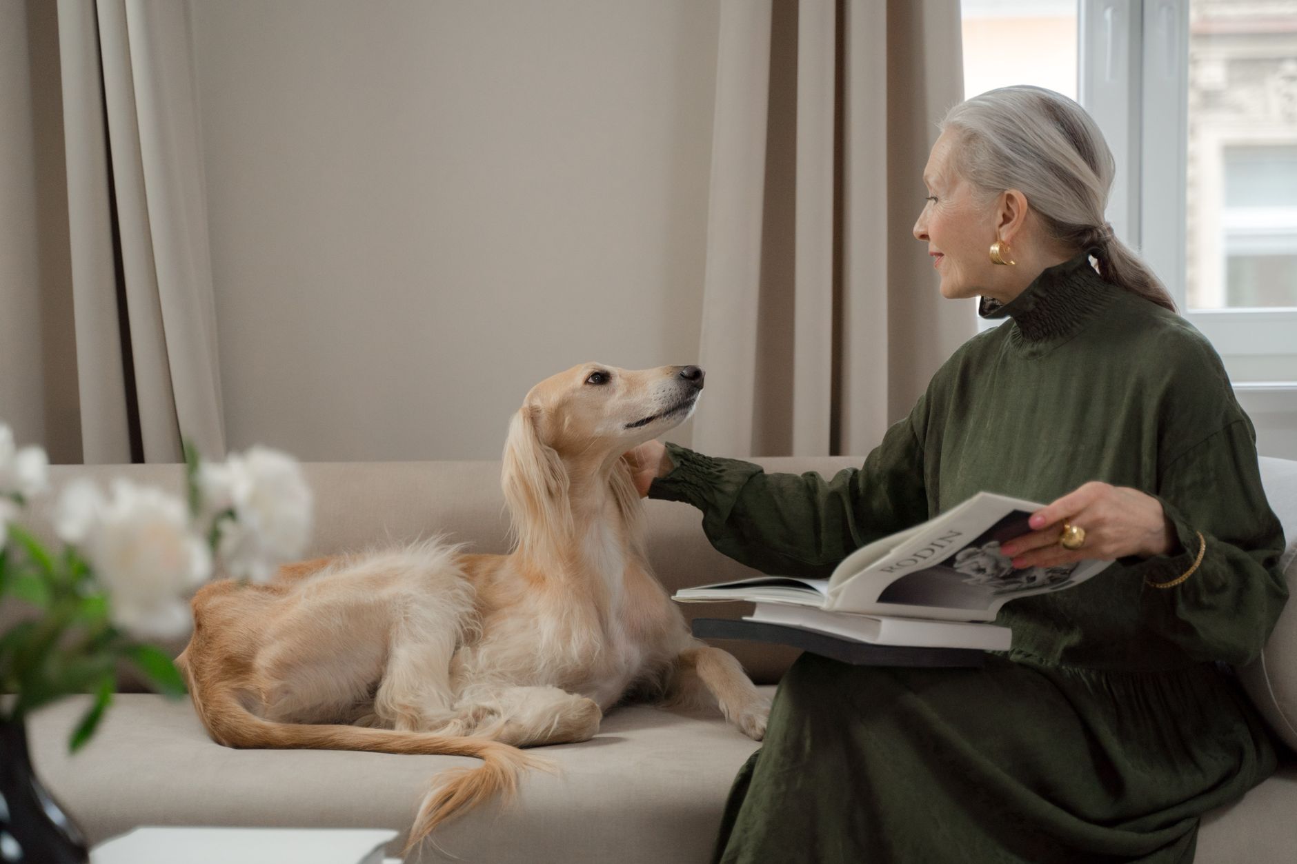 Elegant senior woman reading with a Saluki dog on a living room sofa